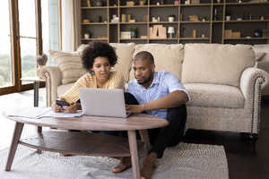 Young couple in their living room, looking at a laptop with bills and a calculator