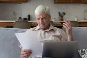 Older man sitting on sofa with laptop and paperwork, looking concerned
